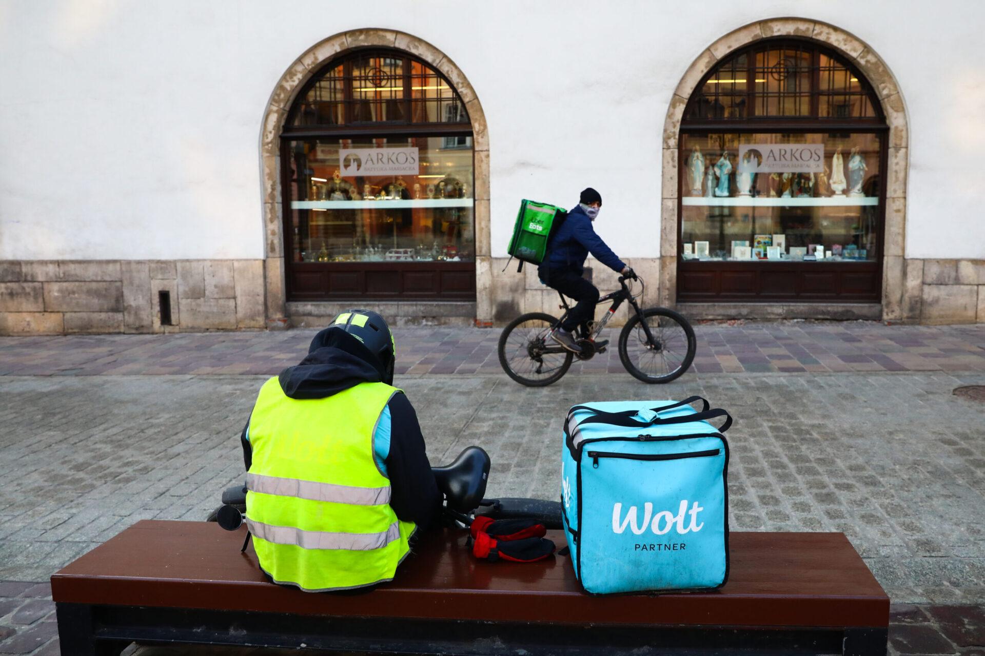 Na pierwszym planie widać kuriera z niebieskim plecakiem na dostawę jedzenia z firmy Wolt, na drugim planie jest kurier, jadący na rowerze, z zielonym plecakiem Uber Eats