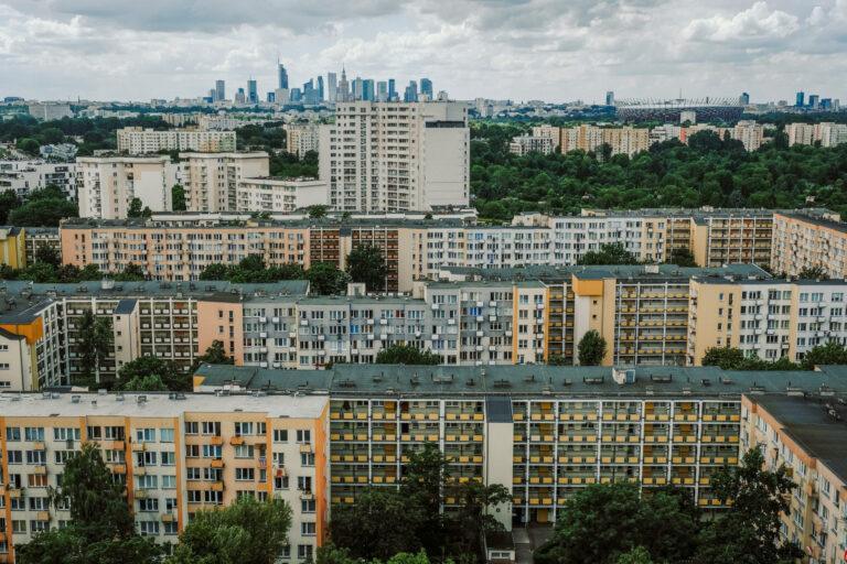 Na pierwszym planie widać bloki mieszkalne, tuż za nimi na horyzoncie widać wieżowce z centrum Warszawy