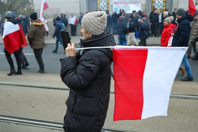 Kobieta trzymająca polską flagę na ramieniu, w tle uczestnicy marszu 11 listopada