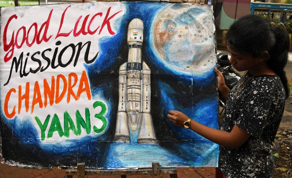 Dziewczynka pokazuje kolorowy plakat z rakietą, księżycem i hasłem: "Good luck mission Chandrayaan3"