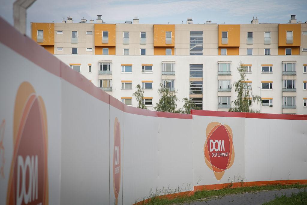 Zdjęcie przedstawia ogrodzenie z logiem Dom Development przed blokiem