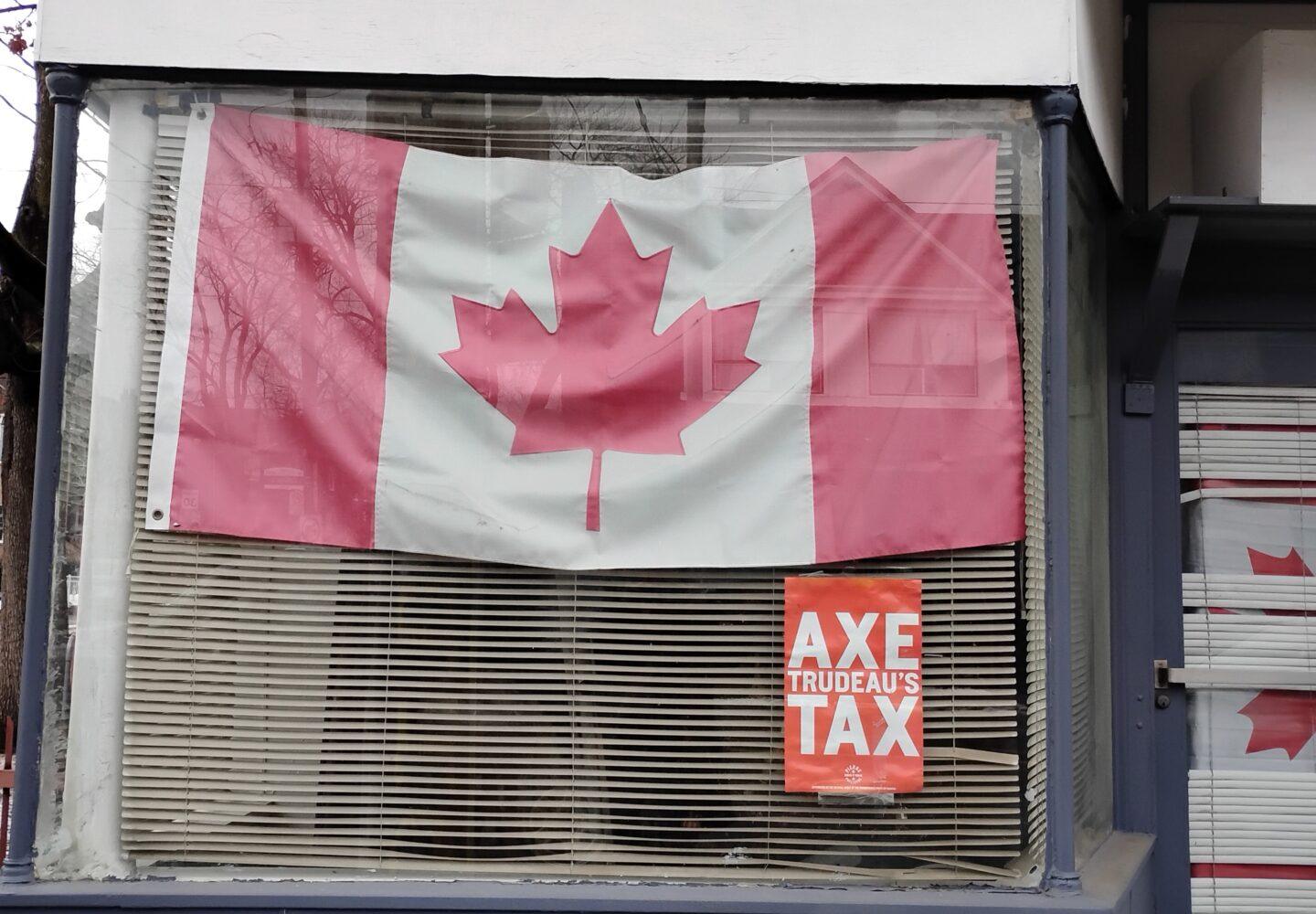 Na zdjęciu widzimy wystawę okienną ozdobioną dużą flagą Kanady oraz plakatem z napisem: „AXE TRUDEAU'S TAX”. Flaga jest lekko pognieciona i przymocowana do szyby, za którą znajdują się poziome żaluzje. Plakat w kolorze pomarańczowym z białymi literami wskazuje na polityczny sprzeciw wobec polityki podatkowej premiera Justina Trudeau. W tle odbijają się drzewa i budynki, co sugeruje, że zdjęcie wykonano w środowisku miejskim.