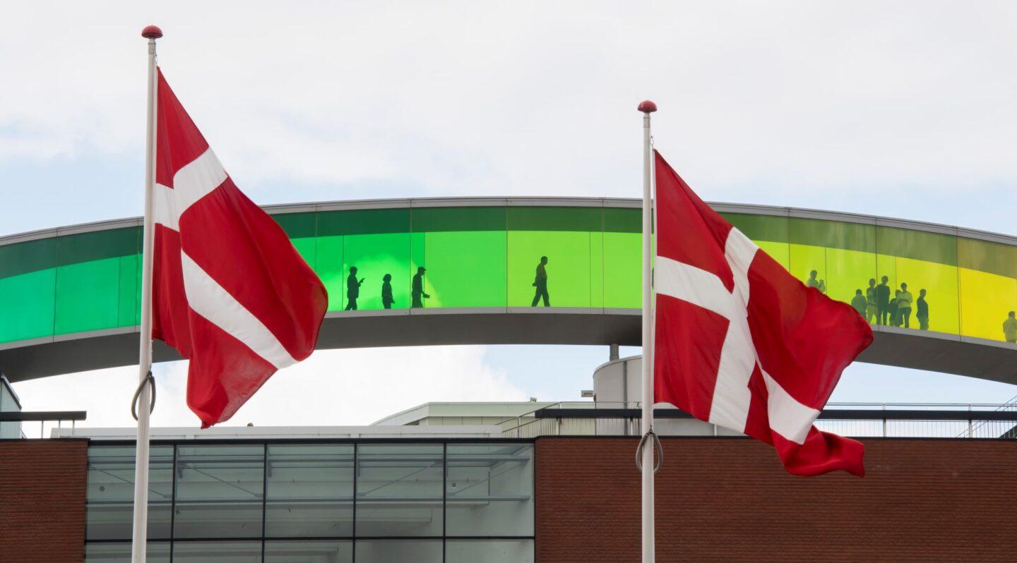 Zdjęcie przedstawia flagę Danii na tle Muzeum ARoS w Aarhus