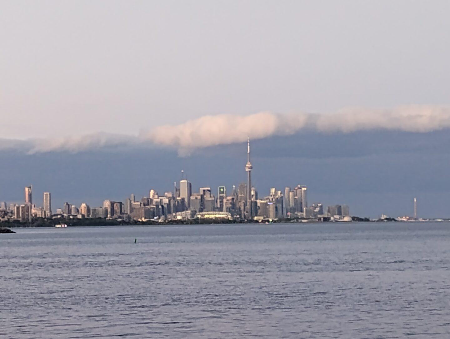Toronto w świetle zachodzącego słońca. Panorama miasta z charakterystyczną wieżą CN Tower na tle dramatycznych chmur i spokojnej tafli jeziora Ontario – obraz metropolii balansującej między naturą a nowoczesnością.