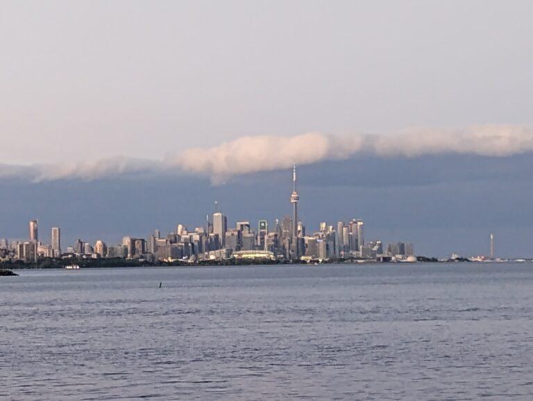 Toronto w świetle zachodzącego słońca. Panorama miasta z charakterystyczną wieżą CN Tower na tle dramatycznych chmur i spokojnej tafli jeziora Ontario – obraz metropolii balansującej między naturą a nowoczesnością.