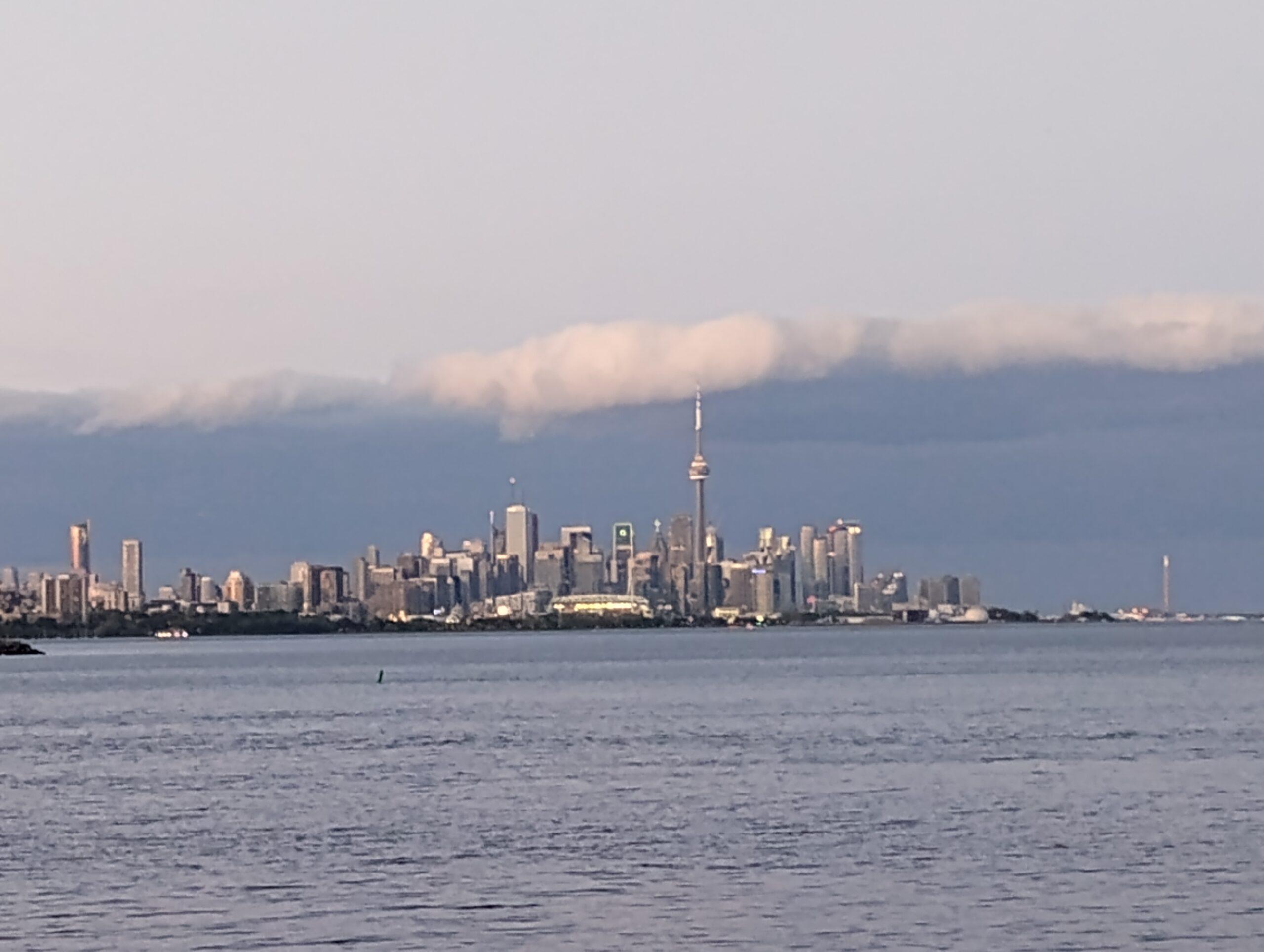 Toronto w świetle zachodzącego słońca. Panorama miasta z charakterystyczną wieżą CN Tower na tle dramatycznych chmur i spokojnej tafli jeziora Ontario – obraz metropolii balansującej między naturą a nowoczesnością.