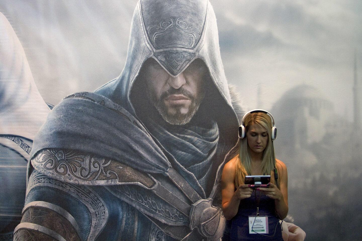 ChatGPT powiedział: Nicole Johnson, przedstawicielka Ubisoft Entertainment, gra w grę wideo stojąc przed reklamą „Assassin's Creed 3” na stoisku firmy podczas targów Electronic Entertainment Expo (E3) w Los Angeles,