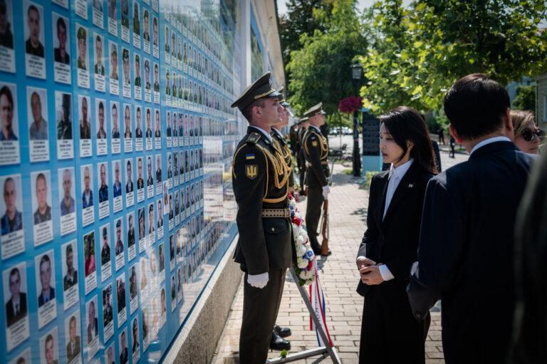 Żołnierze i grupa ludzi pod błękitną tablicą z fotografiami