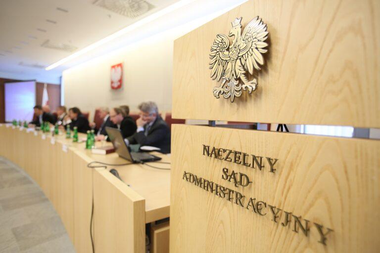 Na pierwszym planie widać godło Polski, a pod nim napis: Naczelny Sąd Administracji. W rozmytym tle widać konferencję prasową z udziałem sędziów