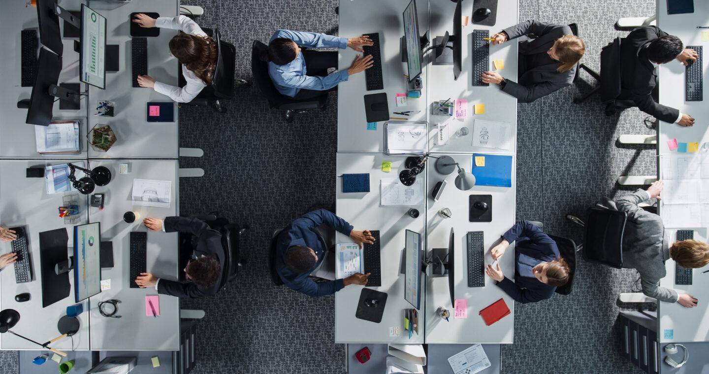 Top Down View of Diverse Colleagues Working In Corporate Office of Hedge Fund. Using Desktop Computers, Discussing Documents, Analyzing Data. Concept of Teamwork and Professional Business Environment
