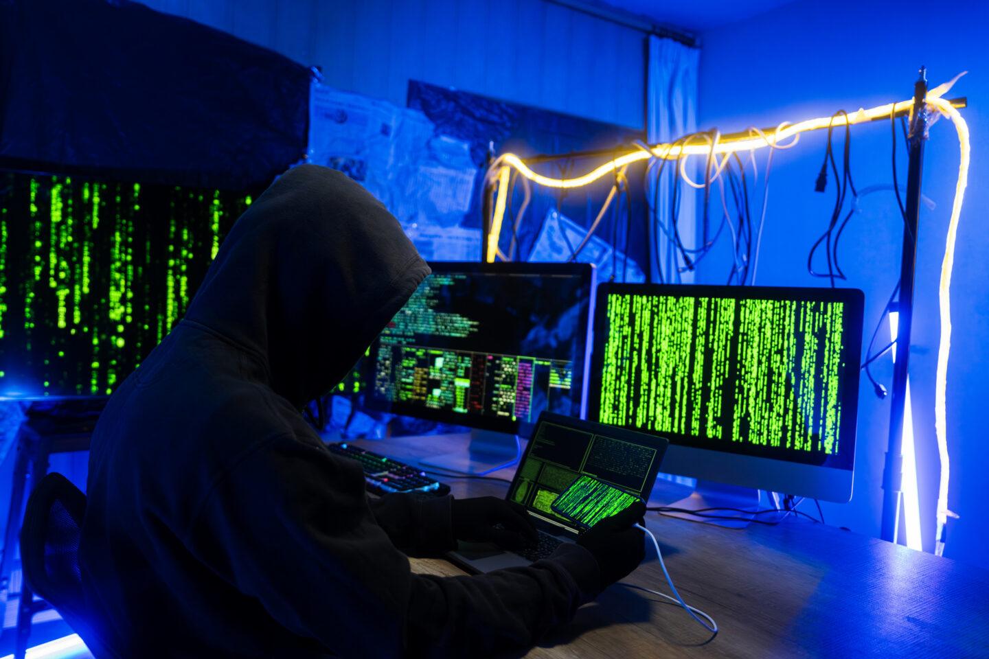Osoba w bluzie z kapturem przed monitorami z widocznym zielonym kodem