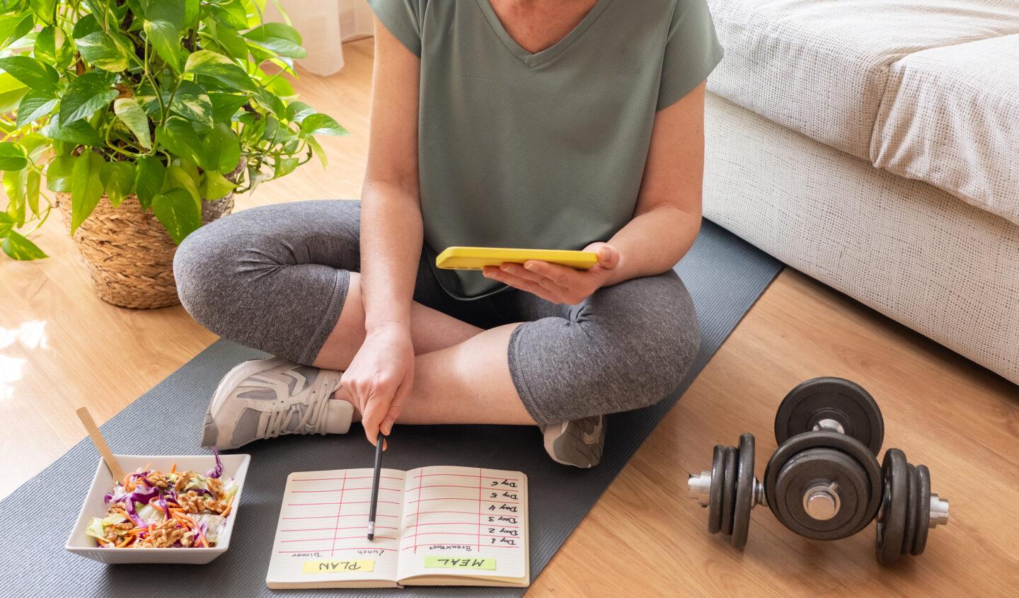 Mature woman sitting on a yoga mat at home, planning her weekly meals and workouts, with a healthy salad and dumbbells nearby, promoting a balanced lifestyle