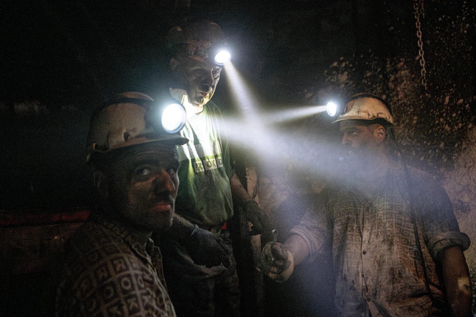 Trzech górników w kaskach z włączonymi lampkami w kopalnianym szybie.