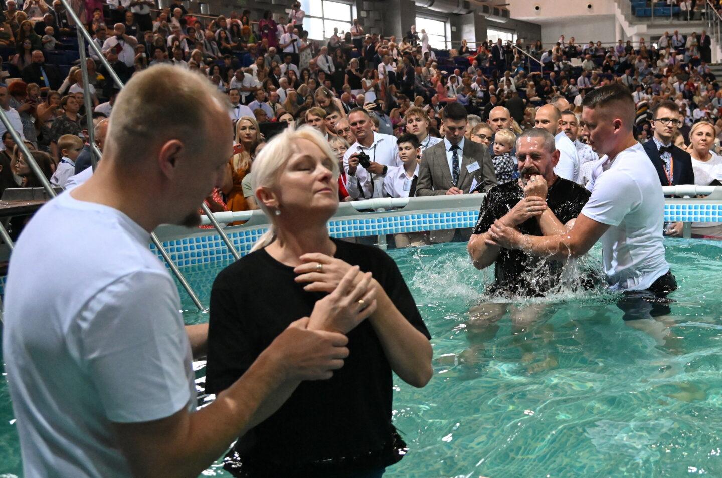 Okrągły basen, a w nim cztery osoby w białych i czarnych koszulkach - ceremonia chrztu nowych członków Świadków Jehowy. W tle trybuny obiektu sportowego wypełnione publicznością.