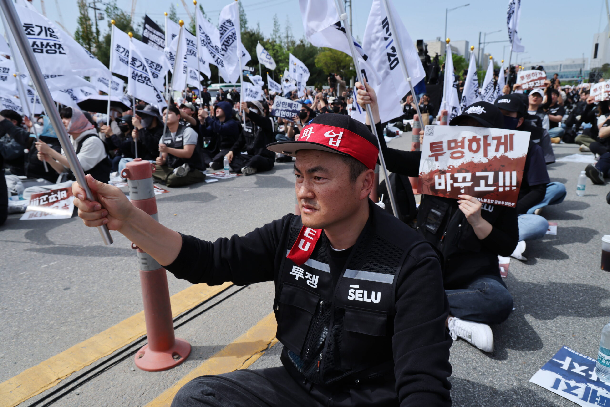 Na pierwszym planie mężczyzna z banerem w języku koreańskim. Na drugim planie grupa osób z flagami z wypisanymi hasłami
