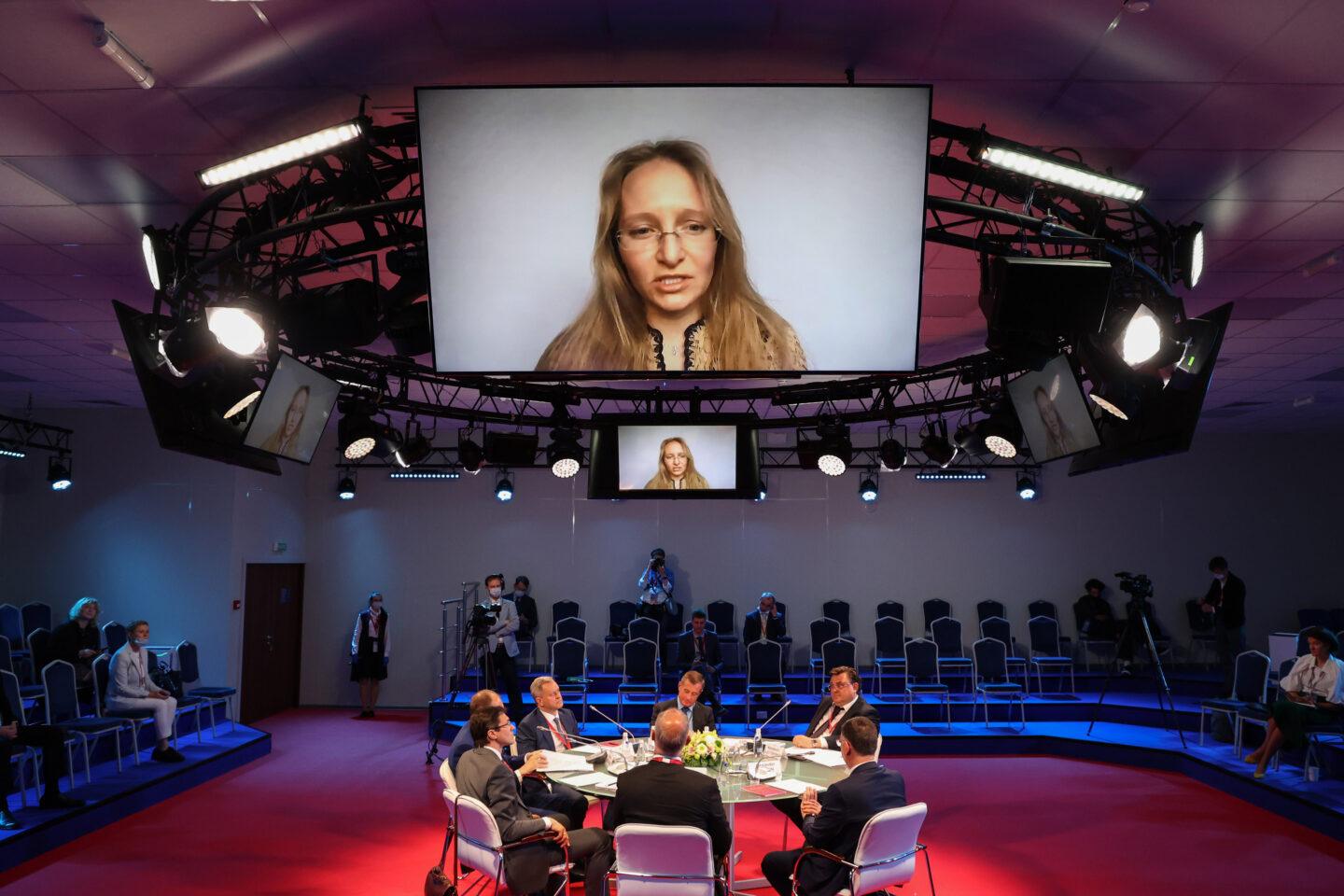 Na wielkim ekranie nad sceną Katerina Tichonowa, córka prezydenta Rosji Władimira Putina; poniżej siedzący przy okrągłym stole debatujący mężczyźni.