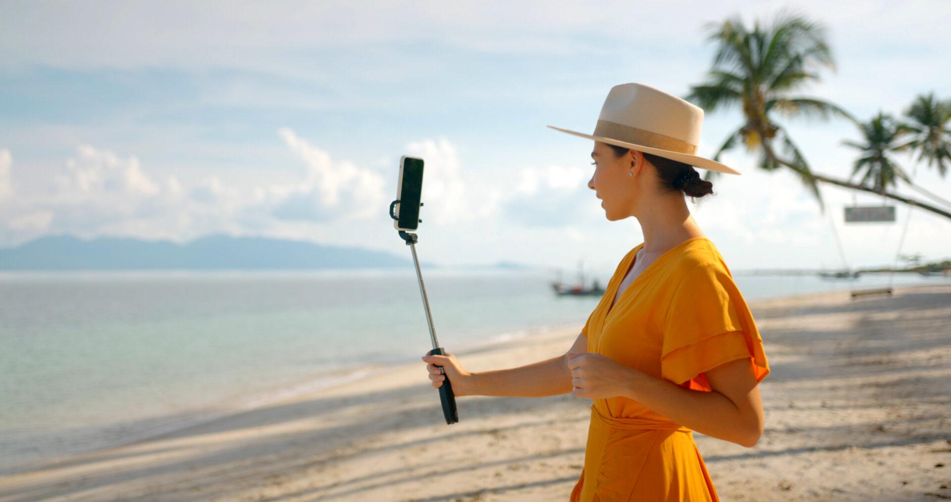 Kobieta w kapeluszu stoi na plaży z telefonem umieszczonym na selfie sticku